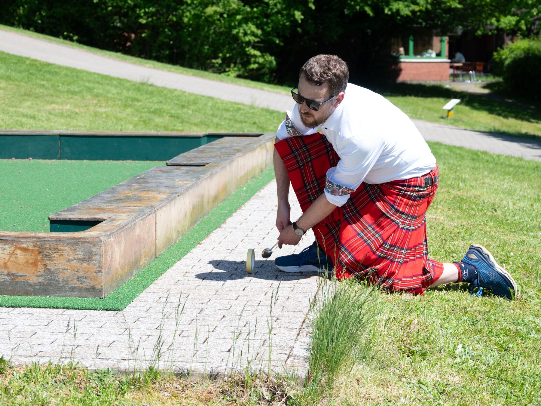 Person in kilt kneeling on pavement and looking around a corner with a Spoon in both hands in front of an upright Quibble to prepare next shot