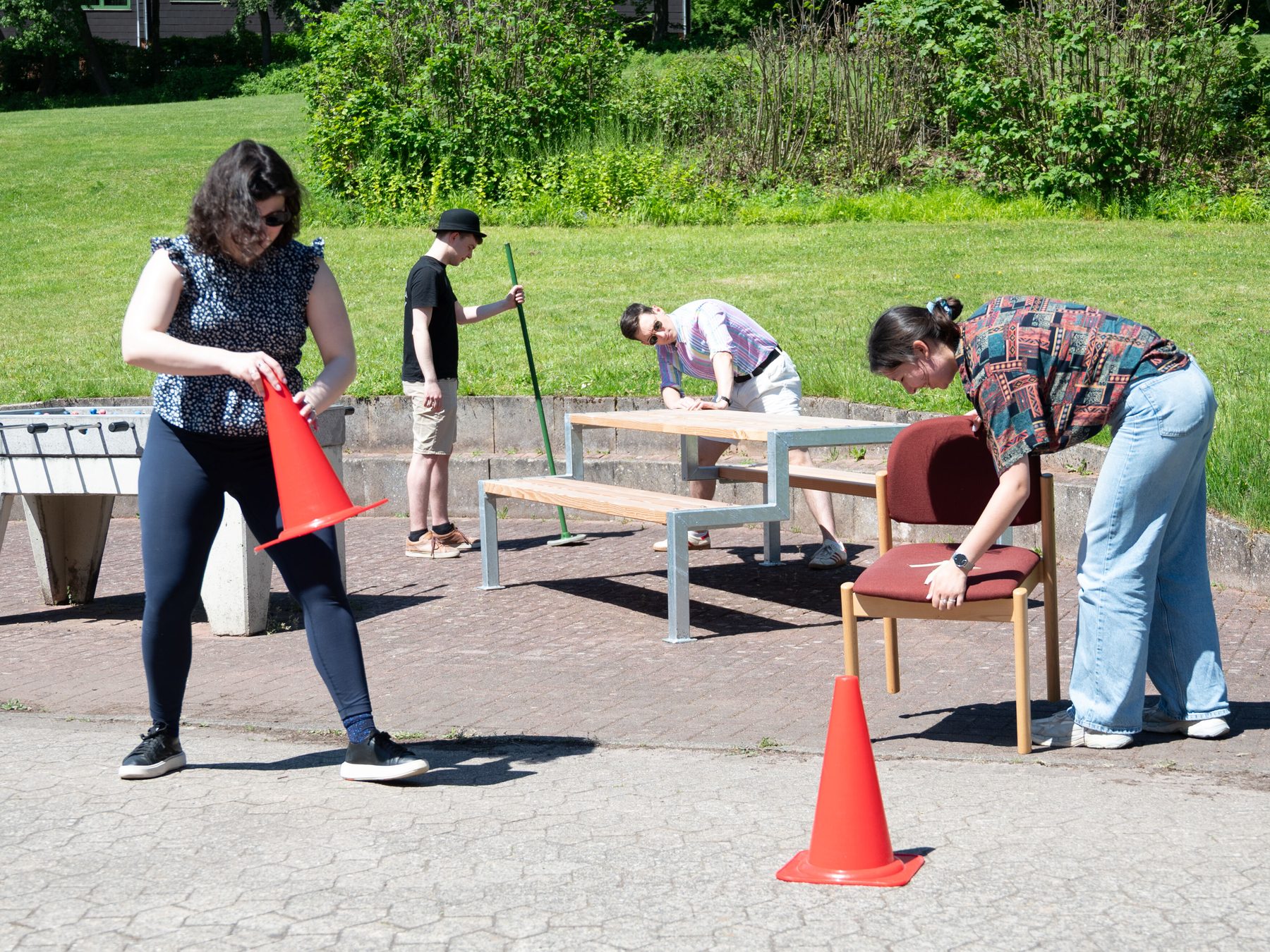 Four people building up a Crolf course by arranging a metal beam, a woode table, a red chair and two pylons on a terrace.